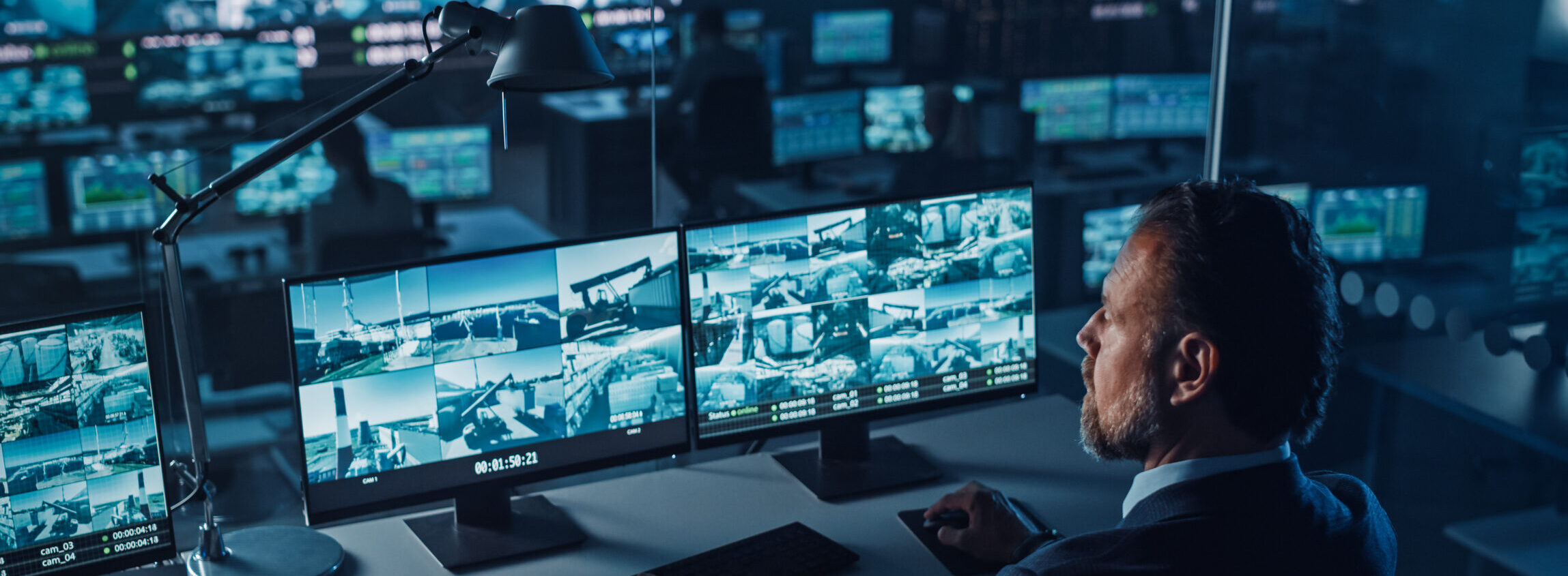 Officer Works on a Computer with Surveillance CCTV Video Footage in a Harbour Monitoring Center with Multiple Cameras on a Big Digital Screen. Employees Sit in Front of Displays with Big Data.